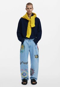 Marineblaues Strickpullover mit gelben Akzenten, getragen über einem Hemd mit Kragen, kombiniert mit hellblauen Jeans, die bunte Tiermotive-Patches aufweisen.