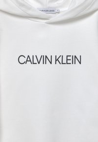 Vit hoodie med en framficka, som har "CALVIN KLEIN" tryckt i fet svart text. Materialet verkar mjukt och lent. Etikett synlig vid halsen.
