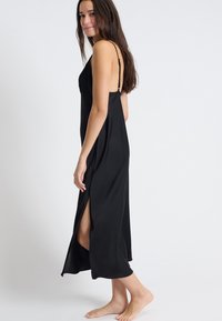 Abito in satin nero con spalline sottili, taglio a metà lunghezza e spacchi laterali per facilitare i movimenti. Texture liscia e design minimalista.