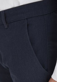 Pantaloni blu scuro con una texture liscia, tasche frontali inclinate e una vita aderente, mostrati con una camicia bianca parzialmente visibile.