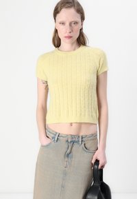 Pulover scurt cu mânecă scurtă din tricot moale galben pal, cu textură ondulată. Asortat cu o fustă din denim deschis la culoare, cu talie înaltă, și cu o geantă neagră.
