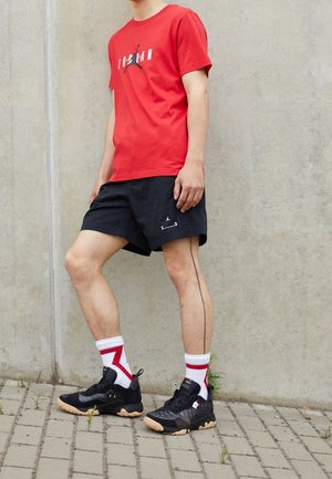 T-shirt en coton rouge avec impression graphique, short noir avec logo, baskets noires, chaussettes rayées blanc-rouge. Fond en béton et pavage.