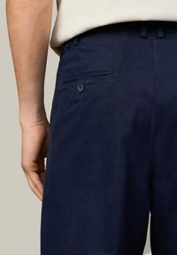 Marineblauwe broek met een gladde textuur, voorzien van een achterzak met knoopsluiting. De taille is gevormd en heeft zichtbare riemlussen.