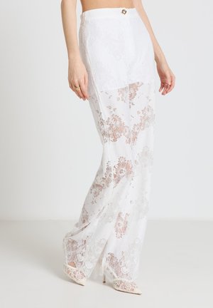 Woman wearing high-waisted white lace pants with floral patterns and white pointed-toe heels, standing against a plain background.