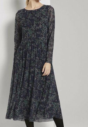 Kvinde iført en mørk, blomsterprint midi-kjole med lange ærmer, samlet talje og gennemsigtige ærmer mod en ensfarvet lys baggrund.
