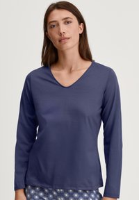 Marineblauwe langemouwen V-hals top gemaakt van zacht materiaal, met een slim fit en een soepele textuur. Hieronder zijn gedeeltelijk gebloemde onderstukken zichtbaar.