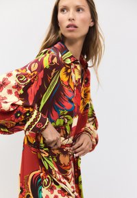 Abito lungo a fiori con colori vivaci rosso, giallo e verde, dotato di scollo con collo e dettagli con bottoni sulle maniche e in vita.