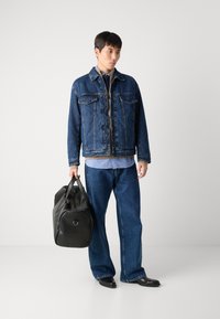 En jeansjacka och vida jeans i mörkblått, lager-på-lager över en ljusblå skjorta. Svart duffelväska med rundad botten och platta handtag.