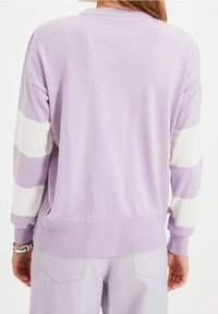 Osoba oblečená v světle fialovém svetru se širokými bílými pruhy na rukávech a odpovídajících světle fialových kalhotách, zobrazená zezadu.