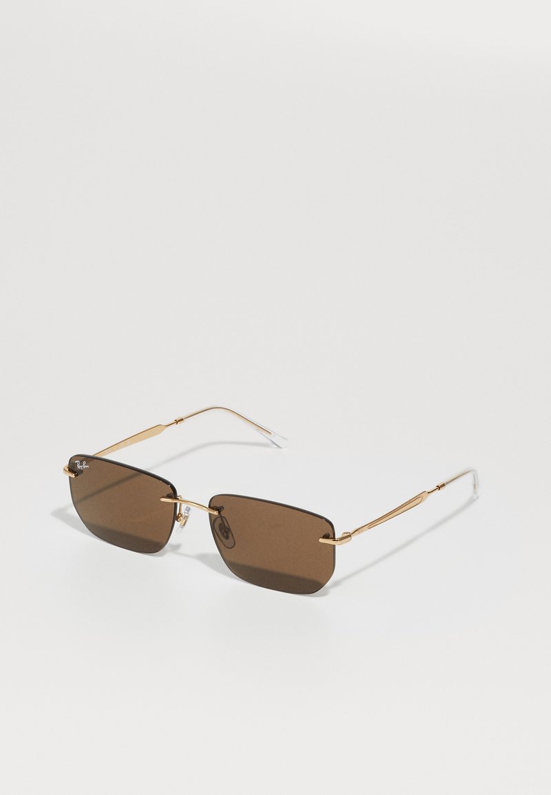 Obdĺžnikové slnečné okuliare Ray-Ban s hnedým nádychom, tenkými zlatými kovovými rámami a priehľadnými plastovými koncami na bielej ploche.