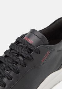 Baskets en cuir noir avec lacets noirs, logo rouge "HUGO" sur la languette et le côté, et semelle blanche, montrées en gros plan.