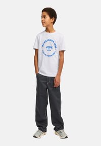 Camiseta de algodón blanca con un logo circular azul en el texto, combinada con pantalones de pierna ancha gris oscuro con costuras en contraste. Las zapatillas completan el atuendo.