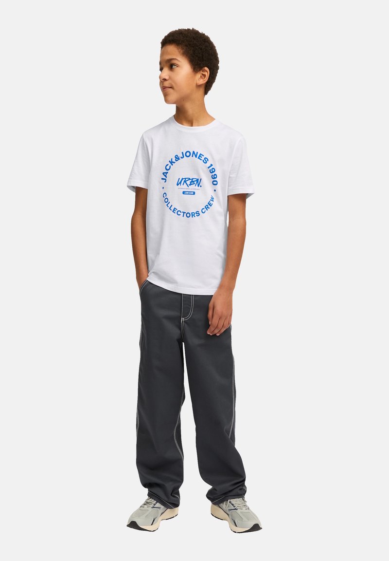 Camiseta de algodón blanca con un logo circular azul en el texto, combinada con pantalones de pierna ancha gris oscuro con costuras en contraste. Las zapatillas completan el atuendo.