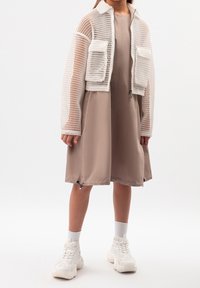 Giacca bianca in rete con due tasche frontali sopra un vestito beige. Sneakers bianche chunky e calze alla caviglia texture completano il look.