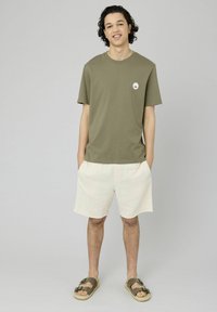 T-shirt vert olive à manches courtes avec un petit logo, associé à un short crème et des sandales marron. Coupe décontractée, tissu en coton doux.