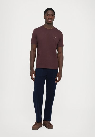 Man staat in een kastanjebruine T-shirt met korte mouwen, marineblauwe broek met grote voorzakken en bruine sneakers tegen een eenduidige witte achtergrond.