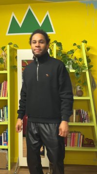 Maglione nero in pile con mezza cerniera e piccolo logo. Abbinato a pantaloni neri lucidi. Sullo sfondo scaffali verdi e pareti gialle.