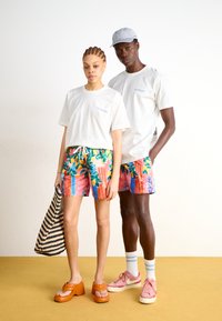 Hvide t-shirts, farverige blomsterprintede badeshorts, orange platformsandaler og pink sneakers. Neutral baggrund med et gult gulv.