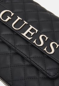 Czarna pikowana torebka z przednią klapą, na której widoczny jest wyraźny srebrny napis "GUESS". Materiał wygląda na fakturowaną skórę z gładkimi akcentami.