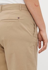 Khakifarbenes Hosen mit glatter Textur, ausgestattet mit einer Gesäßtasche und einem kleinen, mehrfarbigen Logo. Der Bund ist flach mit Gürtelschlaufen.