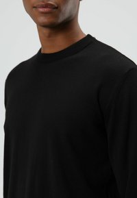 Zwarte trui met een geribde ronde hals en lange mouwen. De stof lijkt zacht met een gladde textuur, en heeft een minimalistisch ontwerp.