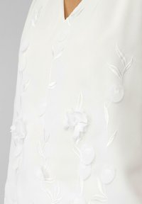 Weißes Kleidungsstück mit V-Ausschnitt, versehen mit strukturierten floralem Stickerei und zarten Blattmustern in passendem Faden, was einen subtilen 3D-Effekt erzeugt.