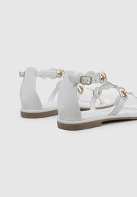 Sandalias de cuero blanco con suela plana, que presentan acentos circulares dorados y una correa ajustable en el tobillo. Diseño abierto con una textura suave.