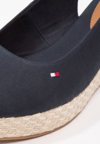 Gros plan d’une chaussure compensée en tissu bleu marine avec une semelle plateforme tissée beige et un petit logo rectangulaire rouge, blanc et bleu.