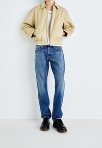 Person klädd i beige zip-up jacka, vit tröja, blå jeans och svarta snörskor, stående mot en enfärgad vit bakgrund.