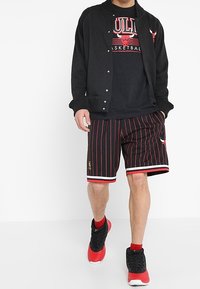 Chaqueta negra con botones a presión sobre una camiseta negra de baloncesto de los Bulls. Pantalones cortos a rayas rojas y negras con borde blanco; zapatillas deportivas rojas y negras.