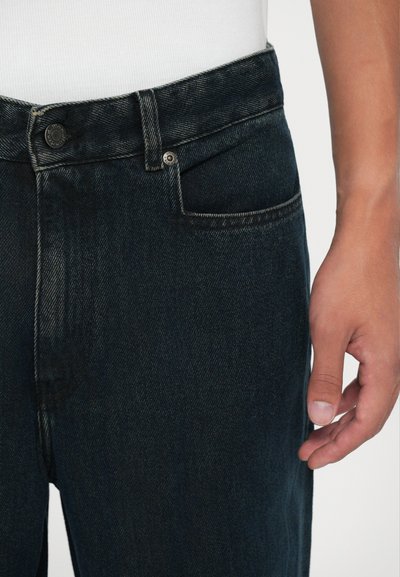 Κοντινό πλάνο της μέσης και της περιοχής της τσέπης ενός σκούρου μπλε τζιν παντελονιού, με ένα ορατό χέρι να ξεκουράζεται δίπλα στο μηρό.