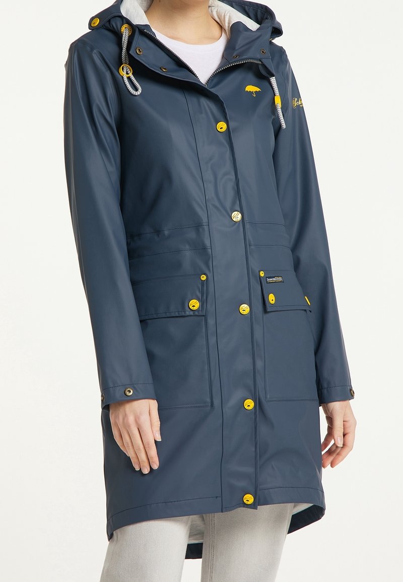 Imperméable bleu marine avec boutons jaunes et logo, poches avant, et capuche avec cordons de serrage, porté sur une chemise blanche.