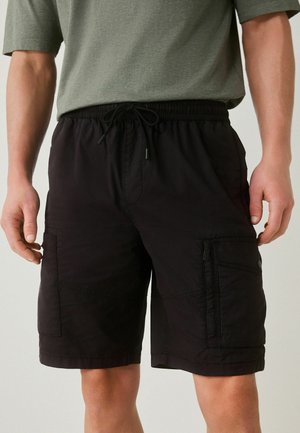 Mees, kes kannab musti lahtisi pükslinaid, millel on nööriga vöökoht ja küljetaskud, koos rohelise lühikese varrukaga särgiga.