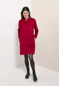 Vestito camicia in cotone a coste rosso con maniche lunghe, colletto con bottoni e tasche laterali. Indossato con collant neri e stivali militari. Texture liscia.