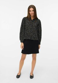 Blusa nera con fiori, maniche a sbuffo lunghe, chiusura con bottoni e texture arricciata, abbinata a una semplice gonna nera e scarpe flat nere.