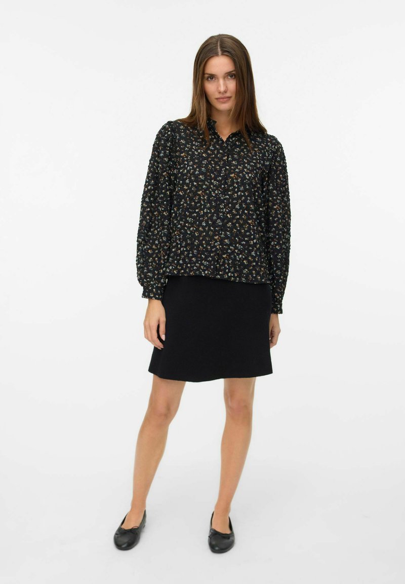 Blusa nera con fiori, maniche a sbuffo lunghe, chiusura con bottoni e texture arricciata, abbinata a una semplice gonna nera e scarpe flat nere.