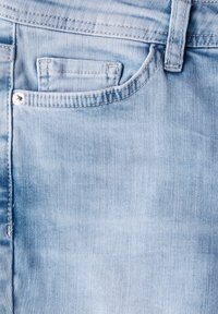 Tissu en denim bleu clair avec texture visible, doté d'une poche avant et d'un accent en métal sur le bouton. Les coutures sont d'une teinte contrastante.
