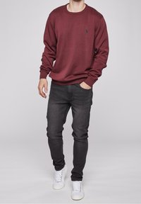 Maroon knitted crewneck sweater with long sleeves, paired with slim-fit dark grey jeans and white sneakers. Subtle logo detail on sweater.