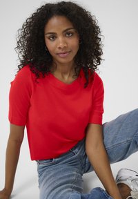 Rode top met korte mouwen en een ronde hals, gecombineerd met lichtblauwe high-waisted jeans. Het model heeft krullend haar en zit ontspannen.