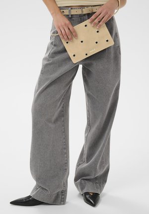 Personne portant un jean gris à jambes larges et des chaussures noires à bouts pointus, tenant une pochette beige avec des œillets métalliques à hauteur de la taille.