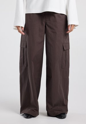 Donkerbruine cargobroek met wijde pijpen en klepzakken aan de zijkant, gedragen met een wit shirt met lange mouwen en zwarte schoenen.