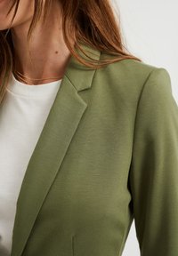 Olivgrön blazer med en strukturerad design, texturerad tyg, slag och en slät vit t-shirt under.