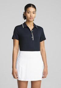 Marineblaues Polohemd mit strukturiertem Kragen, ausgestattet mit weißen Akzenten und Knöpfen. Kombiniert mit einem weißen Sportrock, der einen eleganten, sportlichen Look bietet.