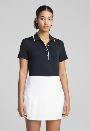 Marinblå polo skjorta med en texturerad krage, som har vita detaljer och knappar. Tillsammans med en vit skort, vilket ger en stilren och sportig look.