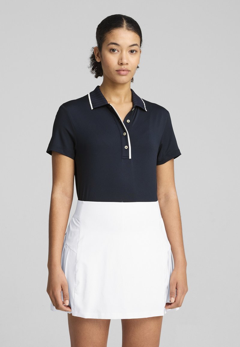 Marineblaues Polohemd mit strukturiertem Kragen, ausgestattet mit weißen Akzenten und Knöpfen. Kombiniert mit einem weißen Sportrock, der einen eleganten, sportlichen Look bietet.