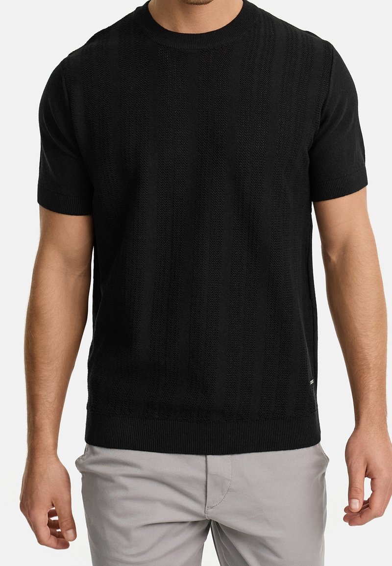 Homme portant un t-shirt en maille texturée noir à manches courtes et un pantalon gris clair, debout les bras détendus le long du corps.