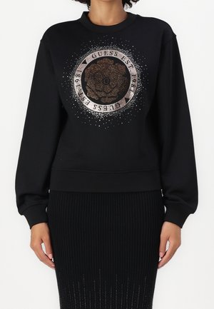 Czarna bluza z dużym okrągłym wzorem róży i tekstem "Guess Est 1981" wykonanym z metalicznych kropek z przodu, noszona z czarną spódnicą.