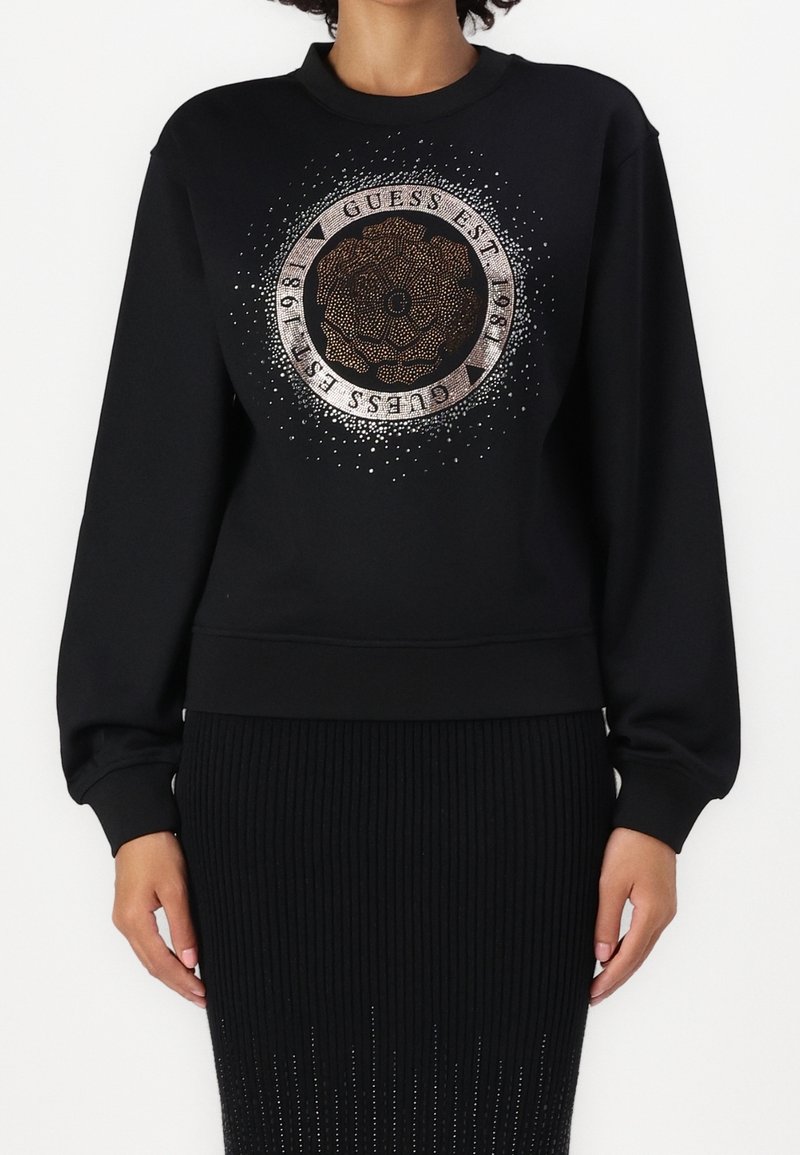 Sweat-shirt noir avec un grand motif circulaire de rose et le texte "Guess Est 1981" en points métalliques sur le devant, porté avec une jupe noire.