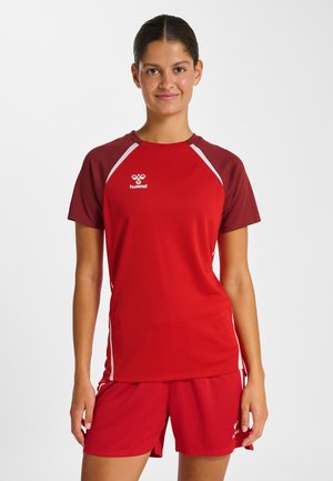 Jeune femme debout, de face, portant un maillot de sport rouge et un short assorti, souriant doucement devant un fond blanc uni.