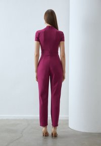 Fuchsia jumpsuit met korte mouwen, aansluitend lijfje en taps toelopende pijpen. Heeft een hoge halslijn en een sleutelgatdetail aan de achterkant.